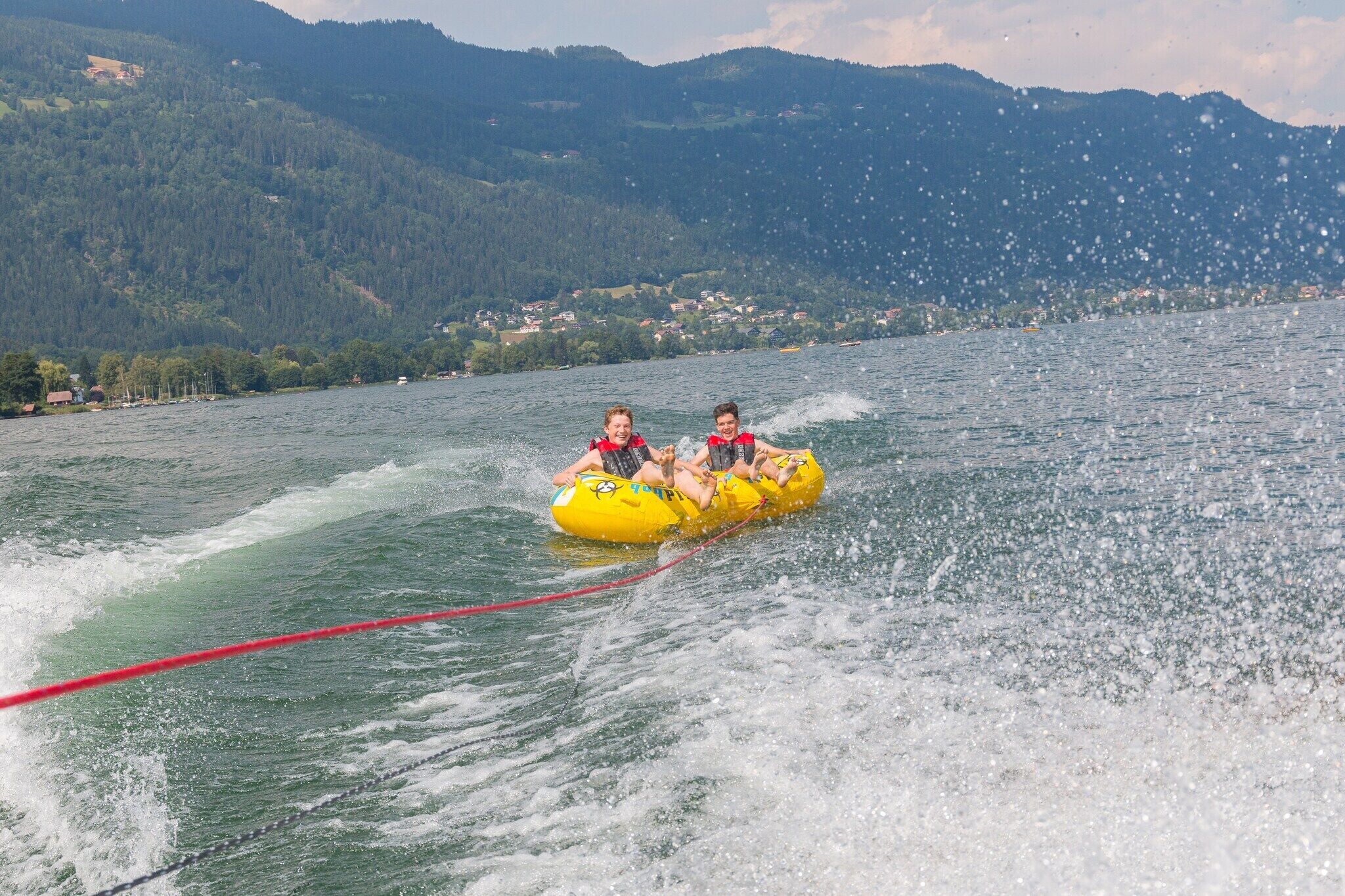 Two people in yellow inflatable tubes being towed on a lake with a red rope, surrounded by mountains and houses in the background.