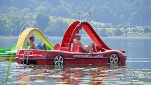 Un bote rojo y amarillo con la inscripción "SPORTSCHUL LASSE" a los costados, flotando sobre el agua con un fondo boscoso.