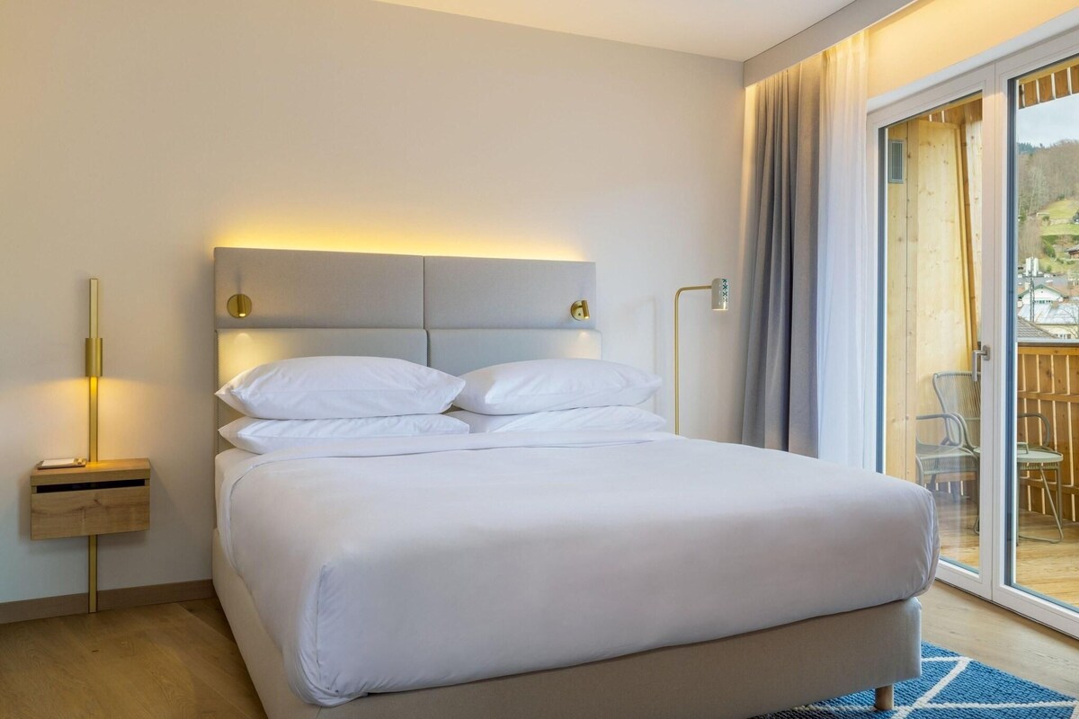 Een hotelkamer met een bed, nachtkastjes, een lamp en uitzicht op de natuur door een glazen deur.