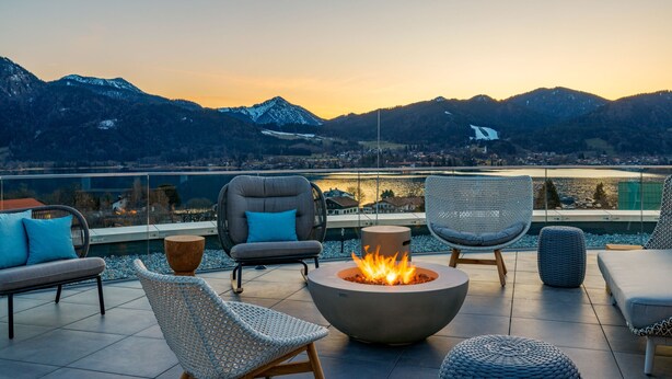 A terrace with outdoor seating, a fire pit, and a view of mountains and a lake. - A-ROSA Tegernsee (Tegernsee)
