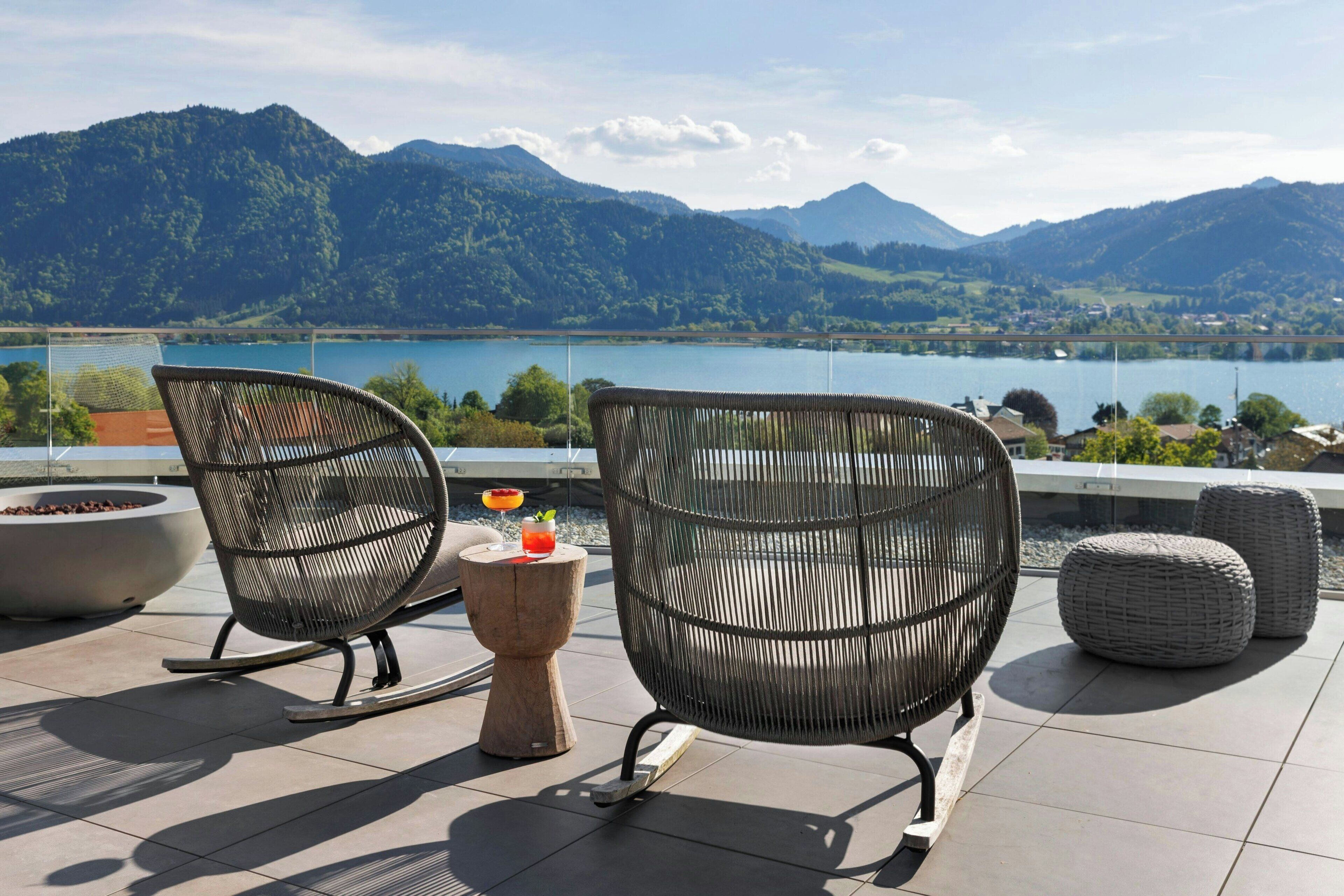 Eine Terrasse mit Korbstühlen und einem Tisch, mit Blick auf einen See und Berge.