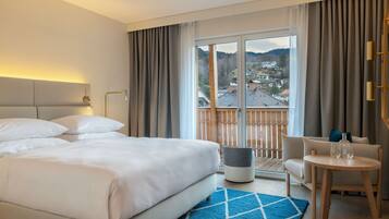 A hotel room with a large bed, a small table with glasses, a chair, and a view of a village through the window.