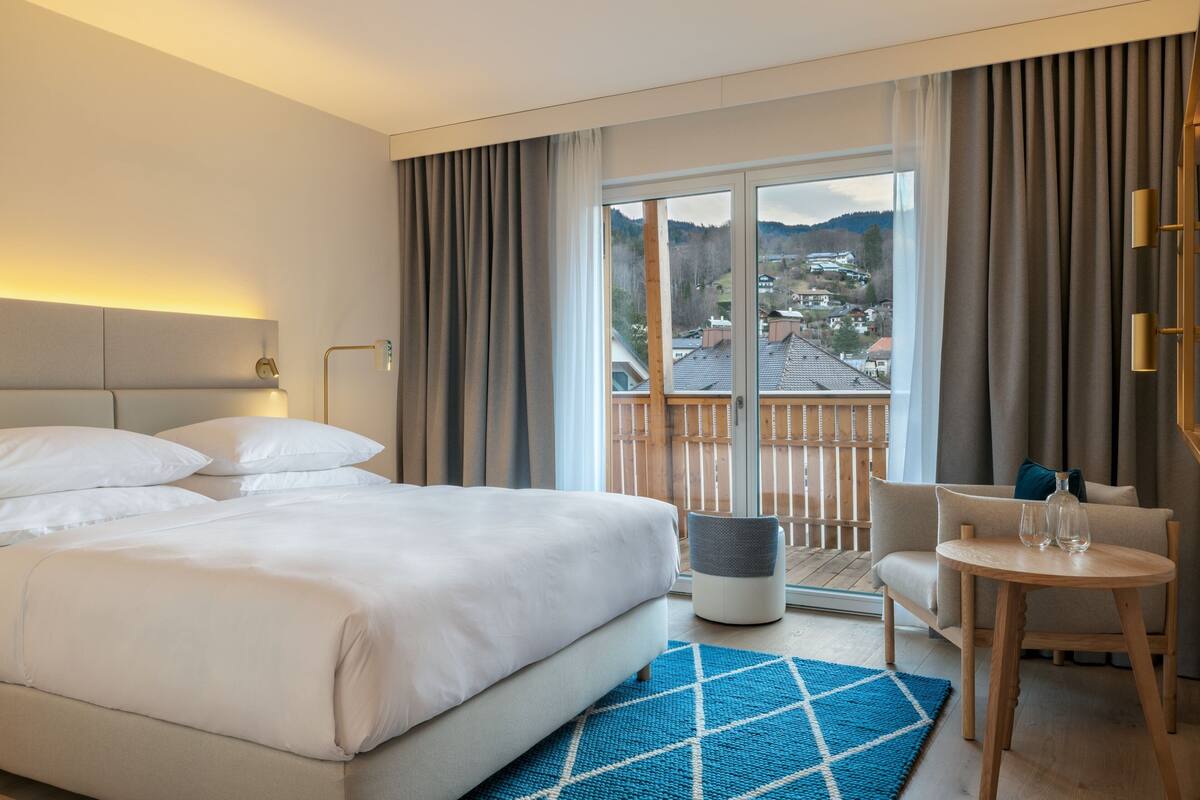 A hotel room with a large bed, a small table with glasses, a sofa, and a view of a town through the window.