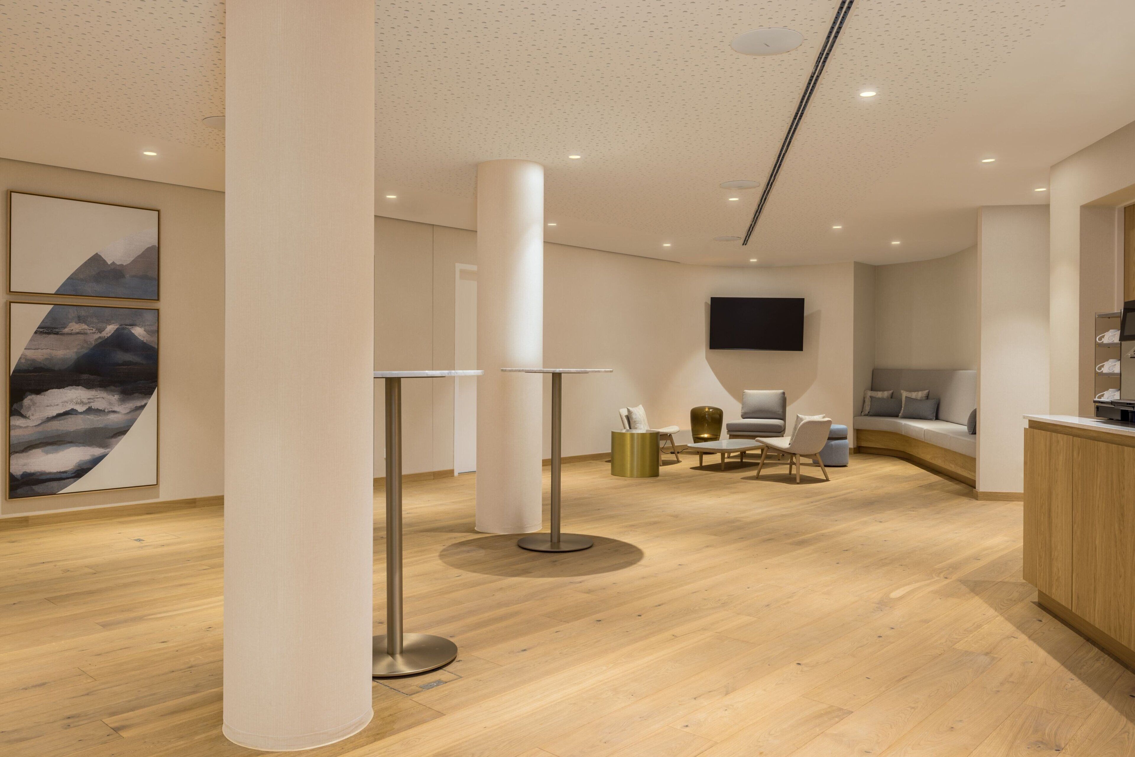 A modern hotel lobby with wooden flooring, a central column, a seating area with a sofa and chairs, and artwork on the walls.