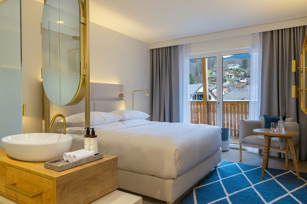 Hotellihuone, jossa on suuri sänky, pyöreä valkoinen pesuallas, puinen meikkipöytä, suuri peili, pieni pyöreä pöytä tuolilla sekä näkymä kaupunkiin suuresta ikkunasta.