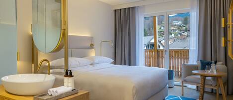 Habitación de hotel con una cama grande, un lavamanos redondo blanco, un tocador de madera, un espejo grande, una mesita redonda con una silla, y vista a un pueblo a través de un ventanal amplio.