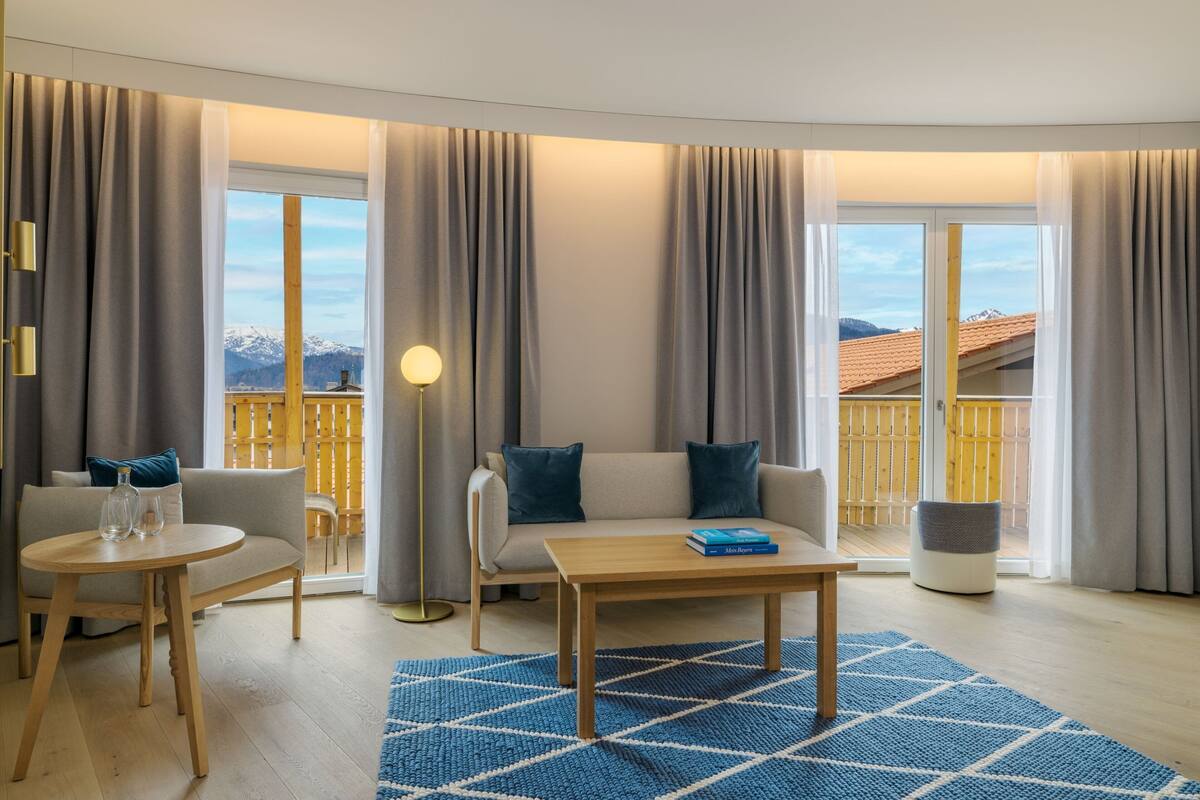 A modern living room with a sofa, a coffee table, and a lamp. There are large windows with curtains, and a view of mountains and a wooden deck outside.