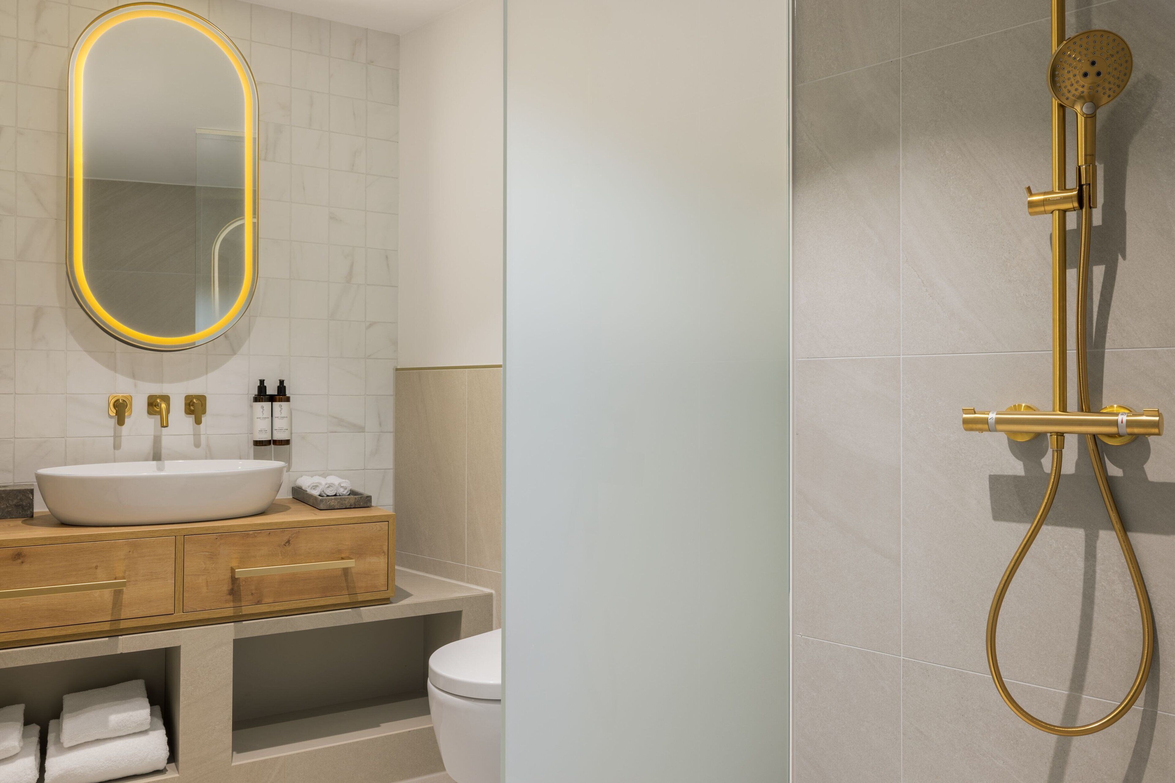Un baño moderno con un espejo ovalado grande, un lavabo blanco, tocador de madera y sistema de ducha dorado.