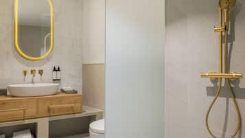 Un baño moderno con un espejo ovalado grande, un lavabo blanco, tocador de madera y sistema de ducha dorado.