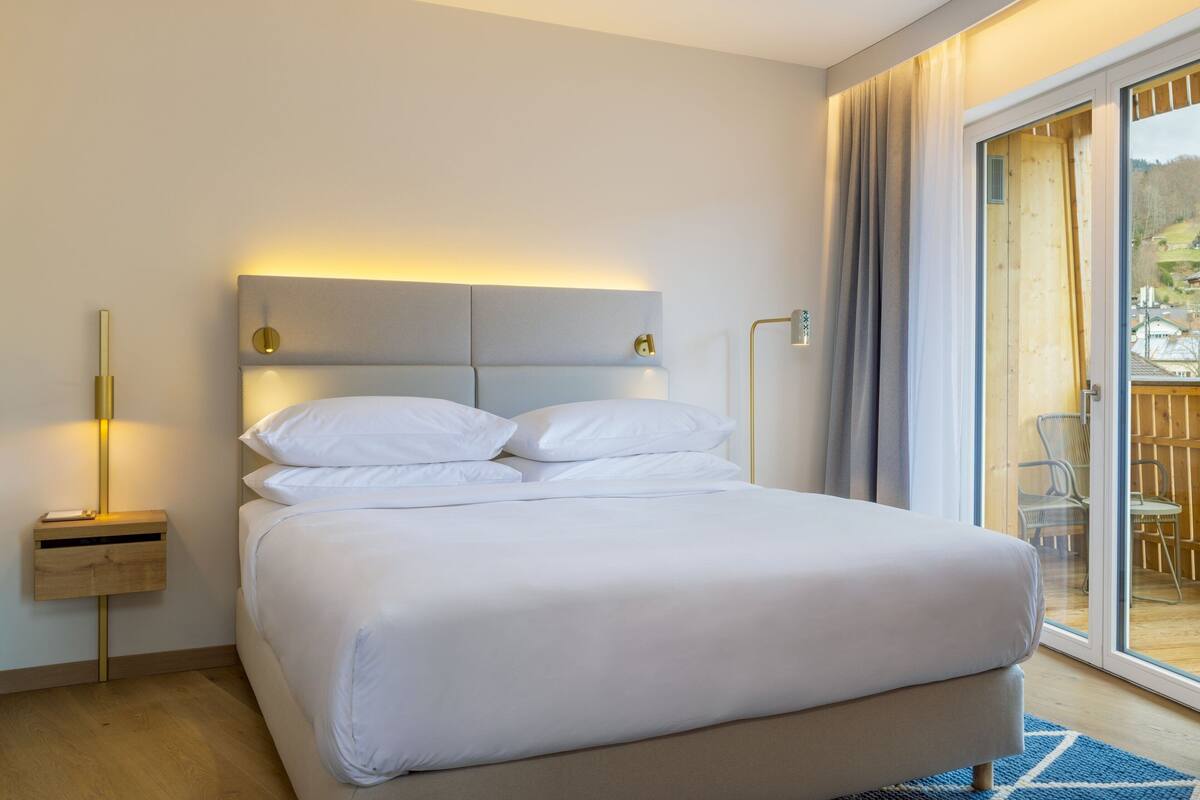A modern hotel room with a large bed, a bedside table with a lamp, and a view of the outdoors through a sliding glass door.