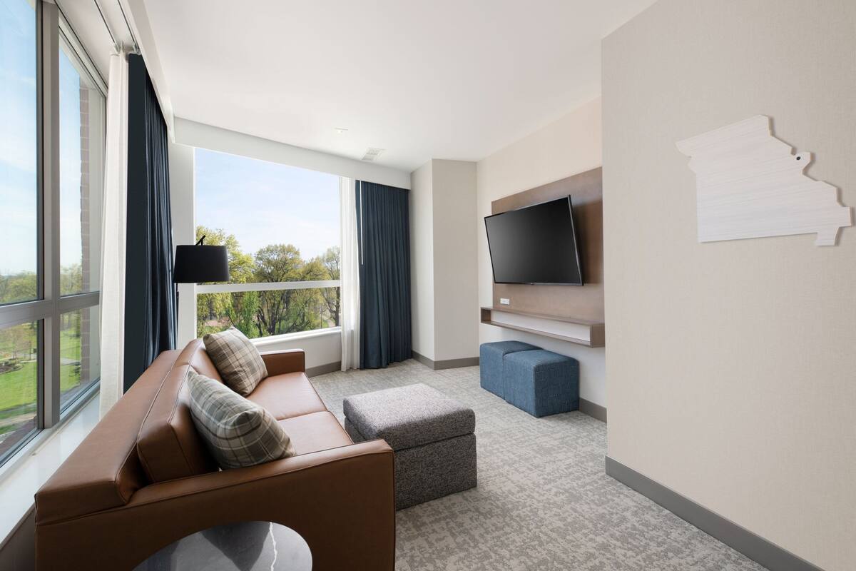 A modern hotel room with a leather sofa, a flat-screen TV, a large window with curtains, and a wall-mounted map.
