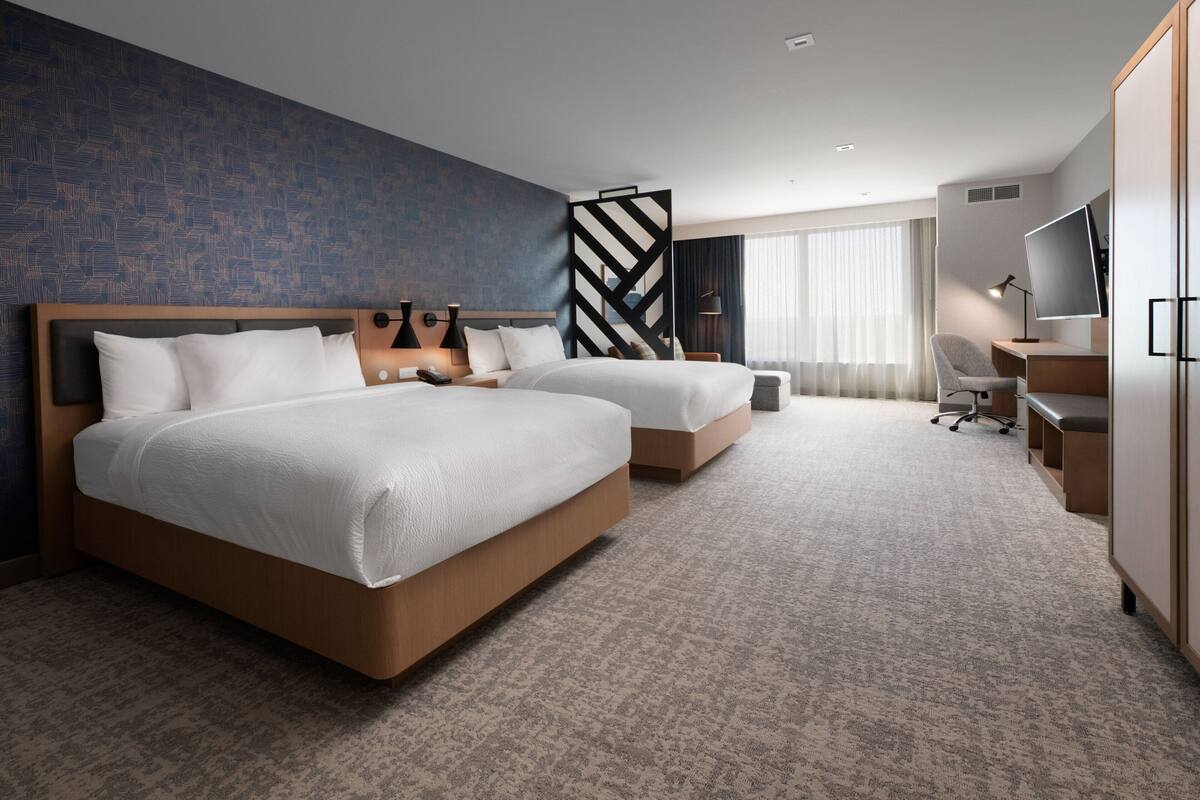 A hotel room with two beds, a desk, a chair, a TV, and a large window.