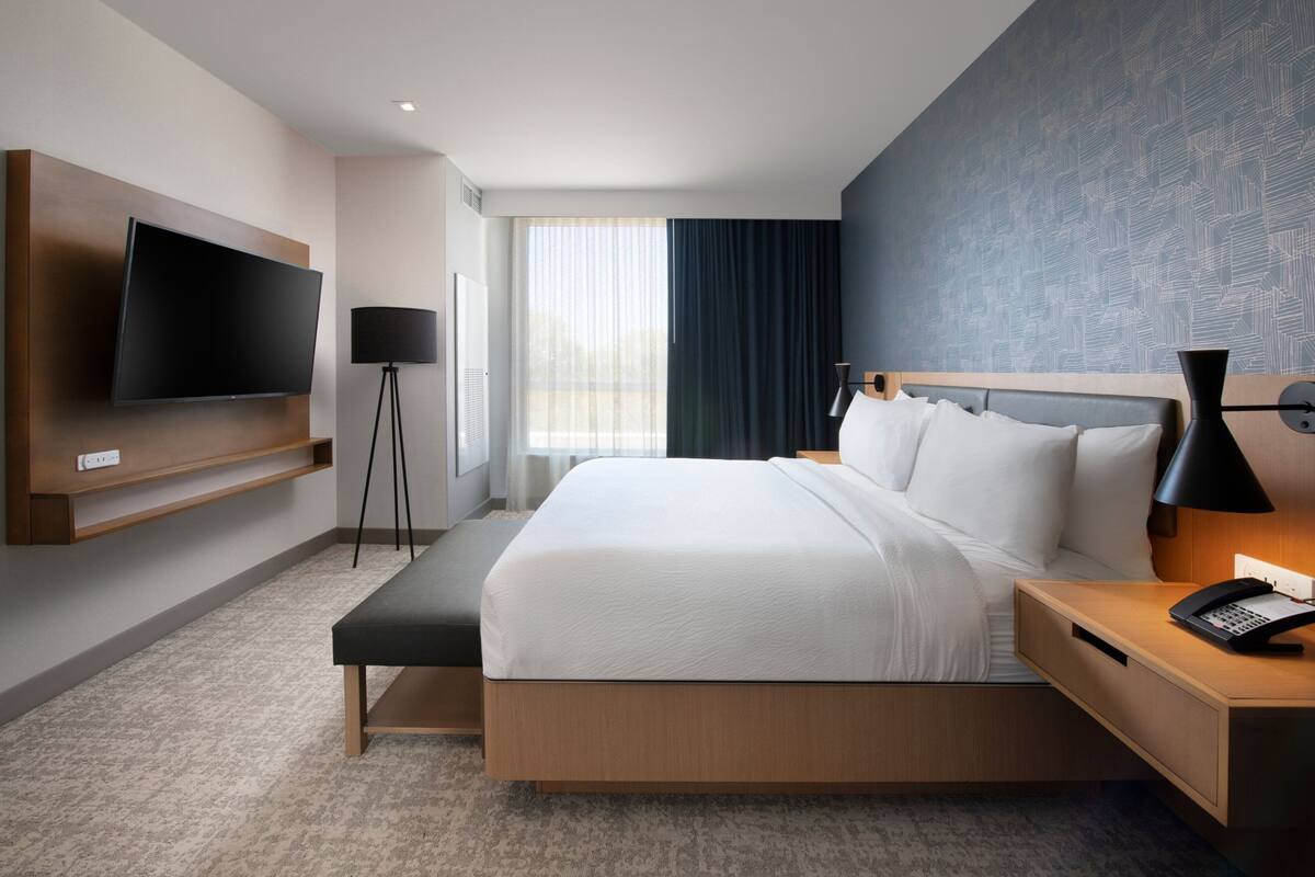 A modern hotel room with a large bed, a flat-screen TV mounted on the wall, a bedside table with a lamp, and a window with curtains.