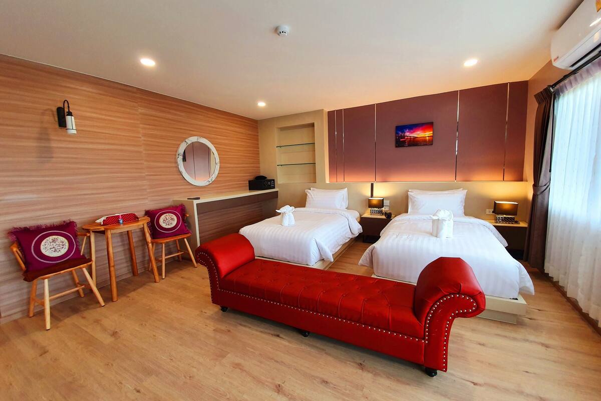 A hotel room with two beds, a red sofa, a wooden floor, a round mirror, and a painting on the wall.