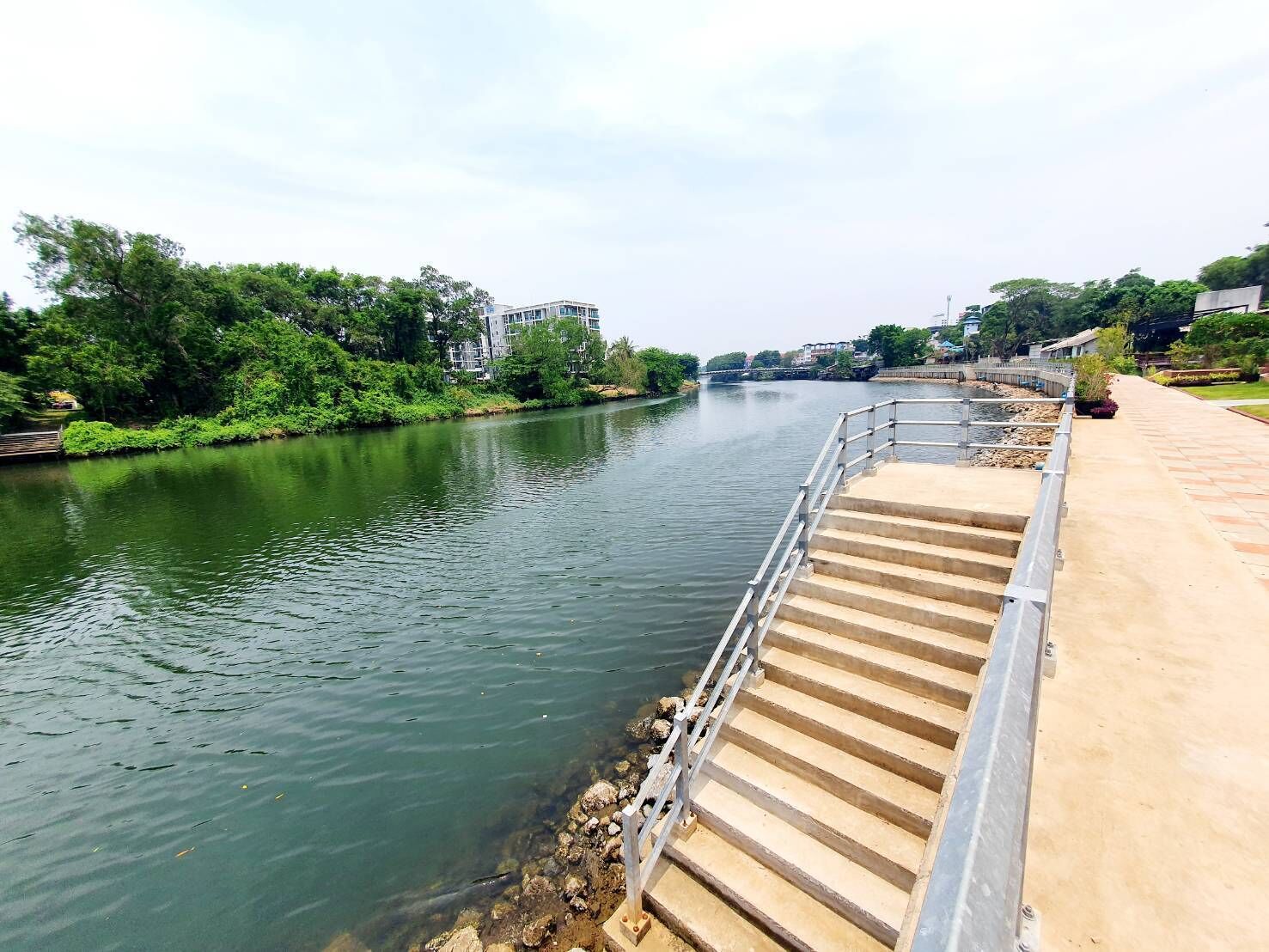 Uma passarela à beira do rio com degraus descendo até a água, cercada por vegetação e edifícios ao fundo.