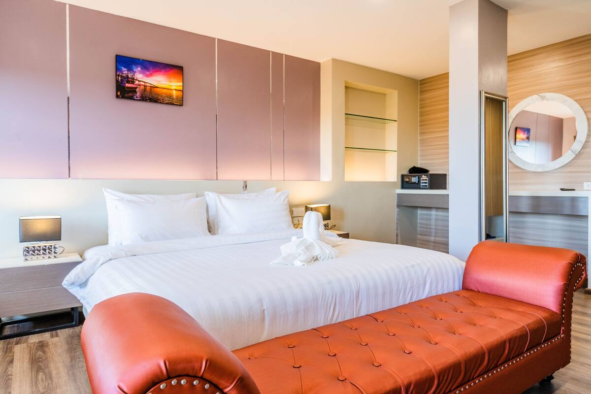 A hotel room with a large bed, a red upholstered bench, a wall-mounted TV, and a painting of a sunset.