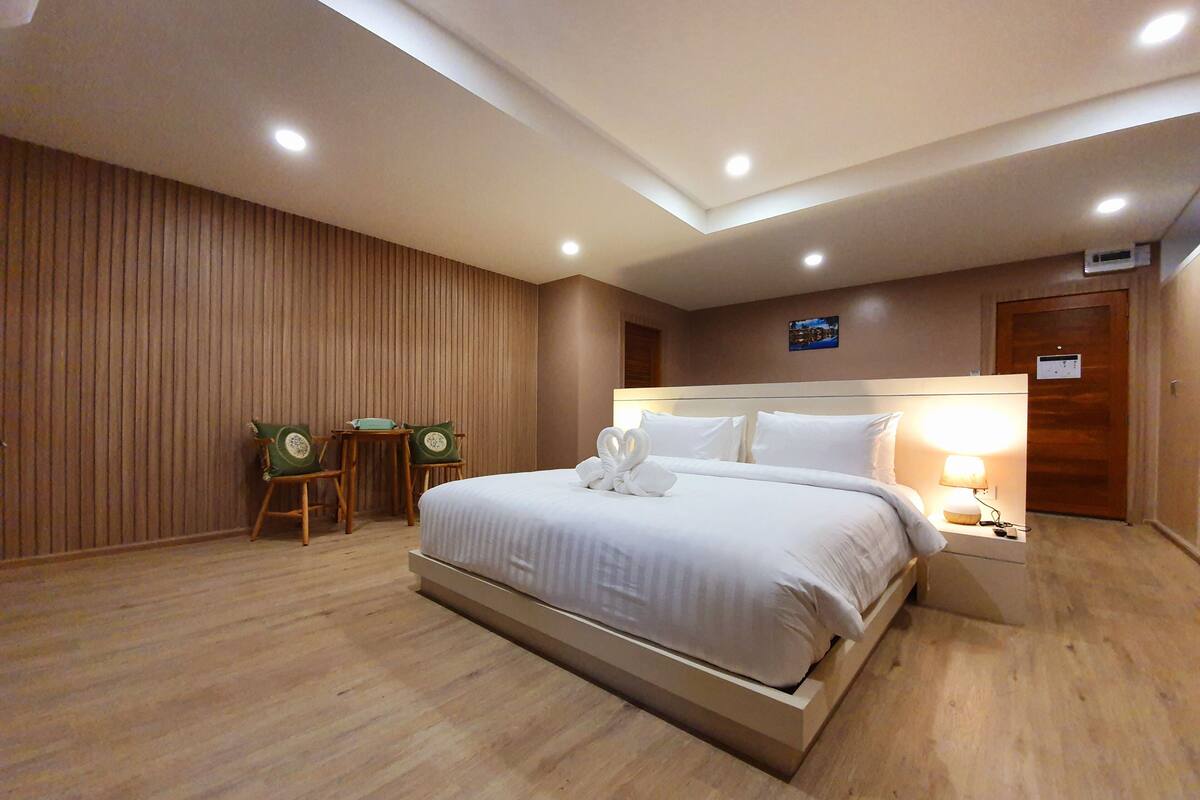 A hotel room with a large bed, a wooden floor, a wooden wall, a small table with chairs, and a nightstand with a lamp.