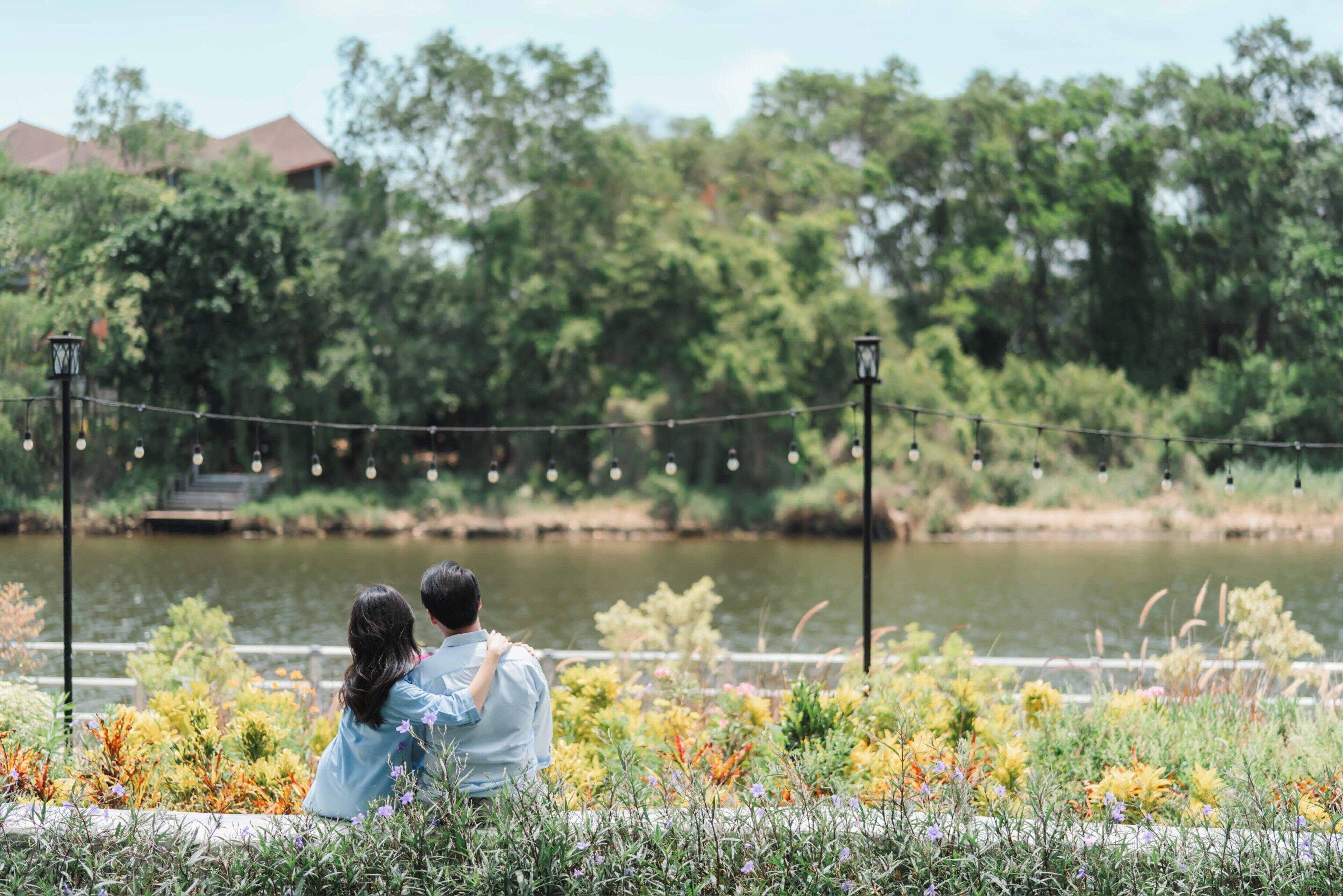 Dos personas sentadas en un banco con vista a un estanque, con diversas flores en primer plano.