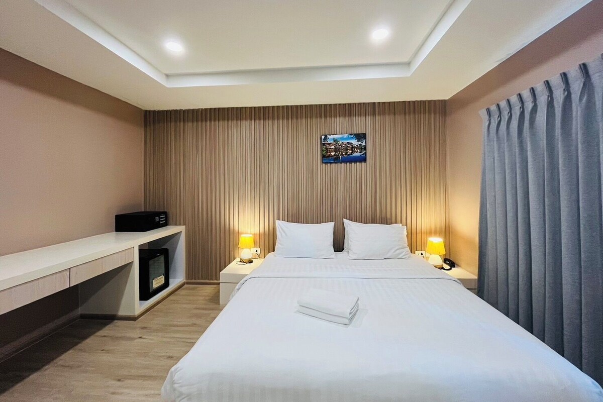 A hotel room with a bed, bedside lamps, a desk with a computer, and a view of a cityscape through a window.