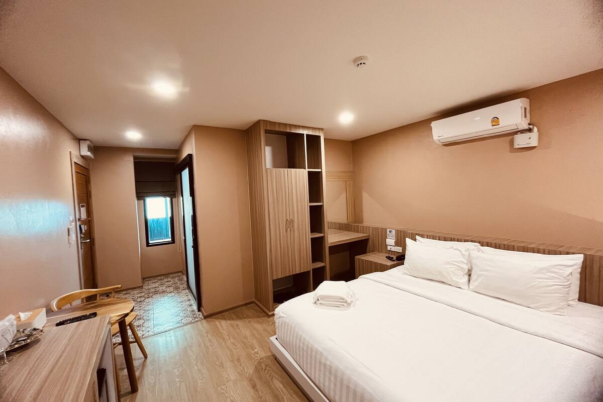 A hotel room with a bed, a desk, a chair, a wardrobe, and an air conditioning unit.