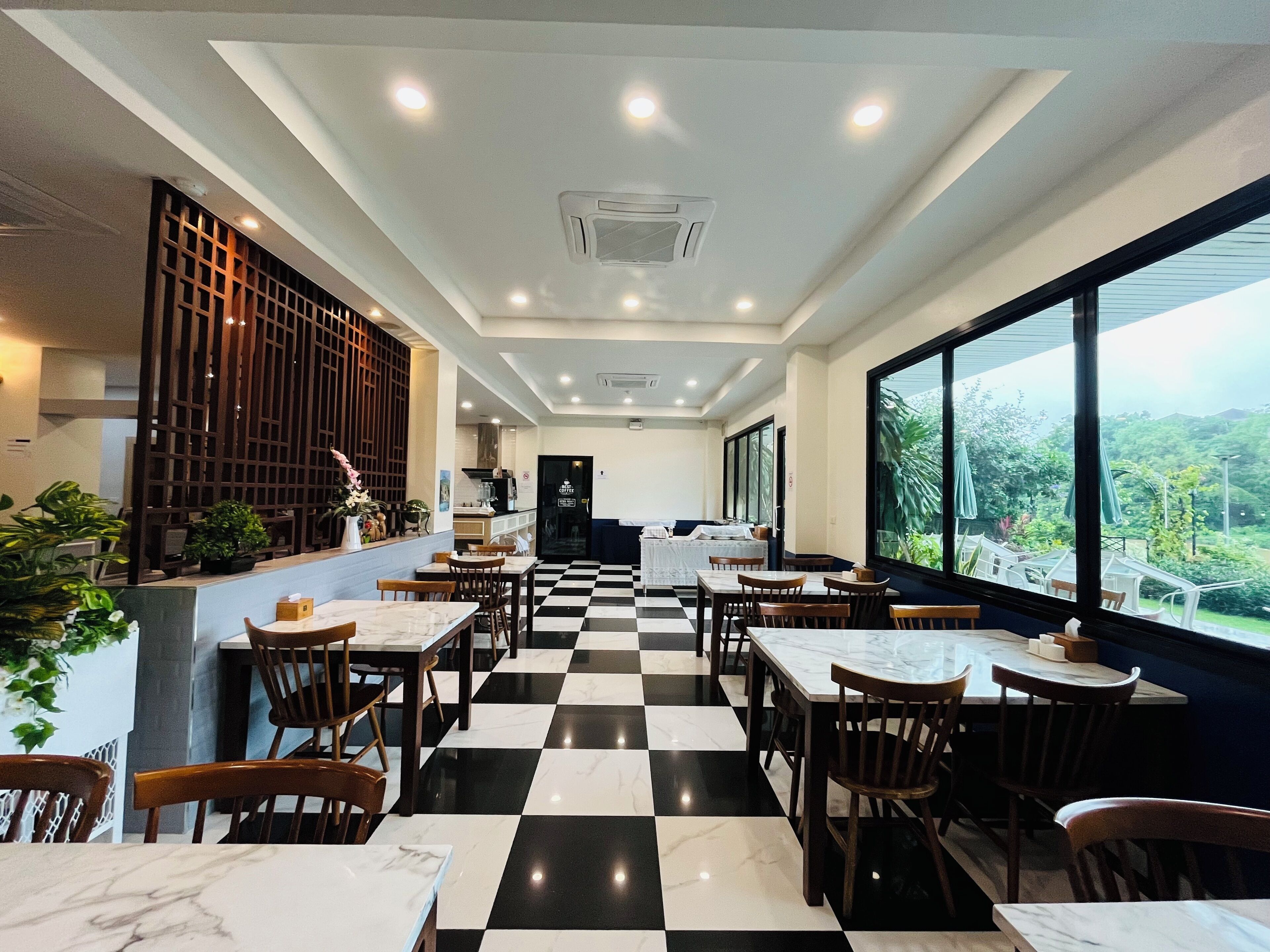 A restaurant with a checkered floor, wooden chairs, and tables. There are potted plants and a view of greenery outside.