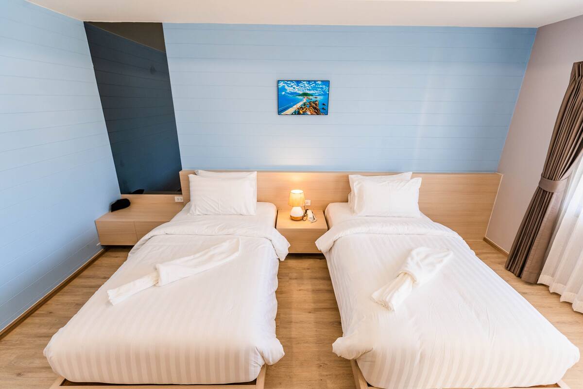 A hotel room with two single beds, a nightstand with a lamp, a painting on the wall, and a wooden floor.