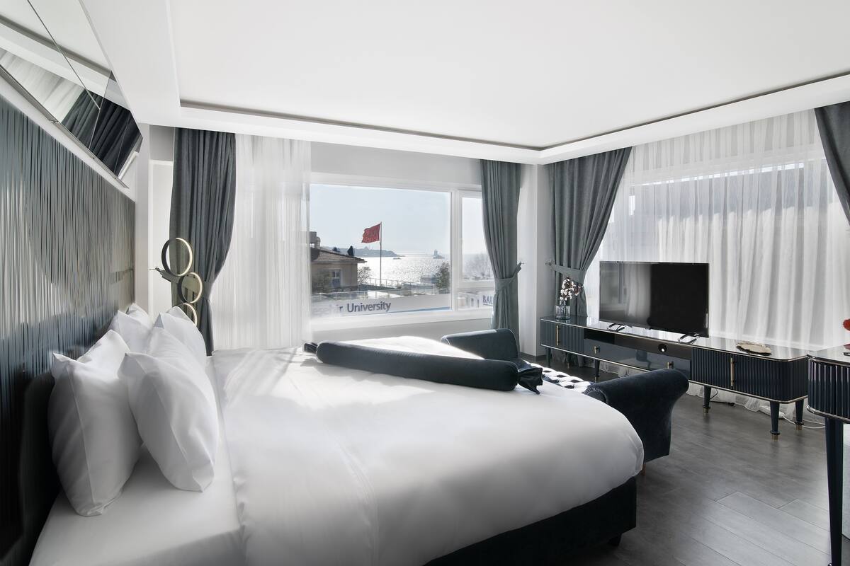 A modern hotel room with a large bed, a desk, a television, and a view of a body of water and buildings.