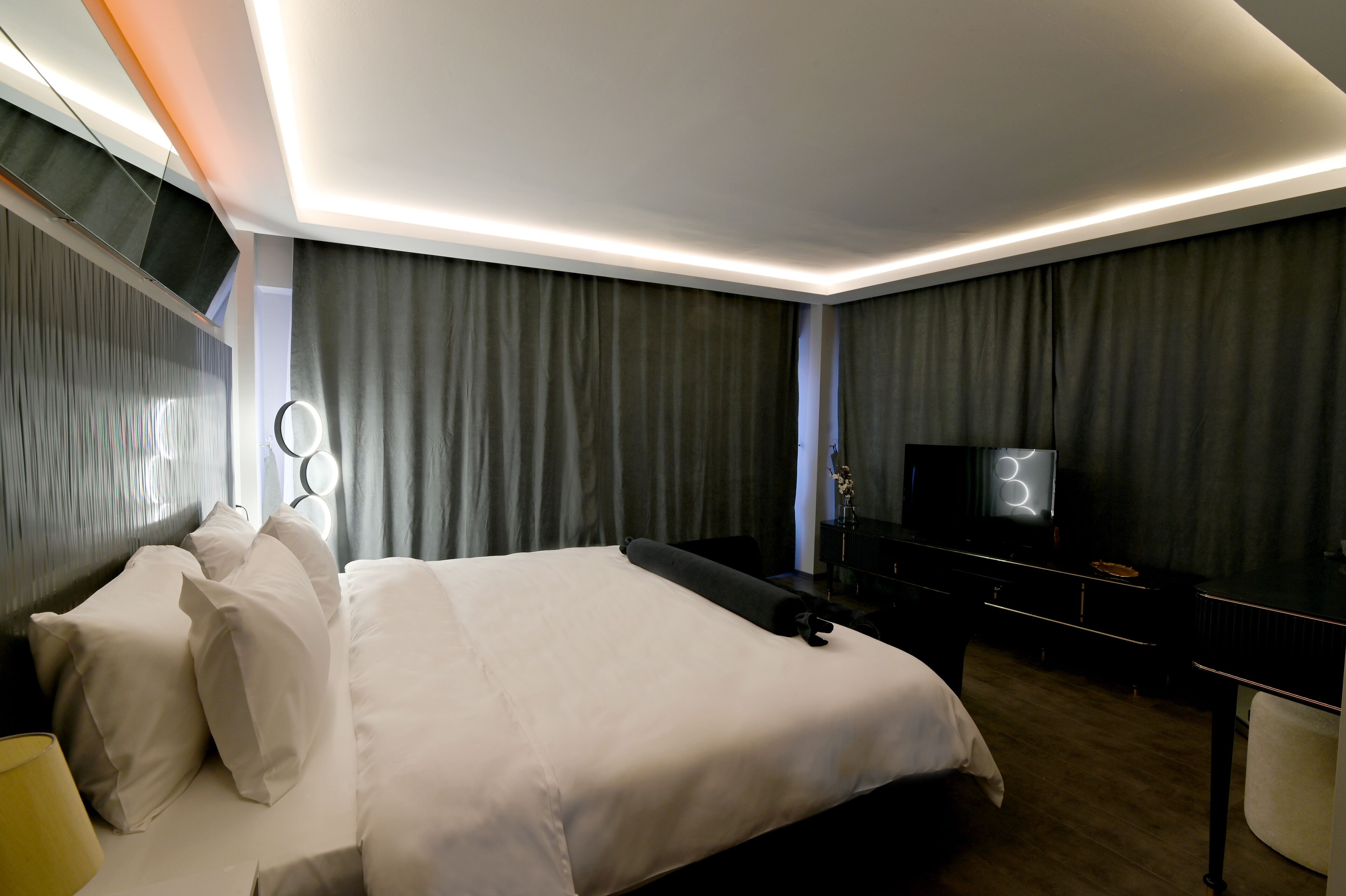 A modern hotel room with a neatly made bed, a flat-screen TV, a vanity with a mirror, and a dark-colored dresser.
