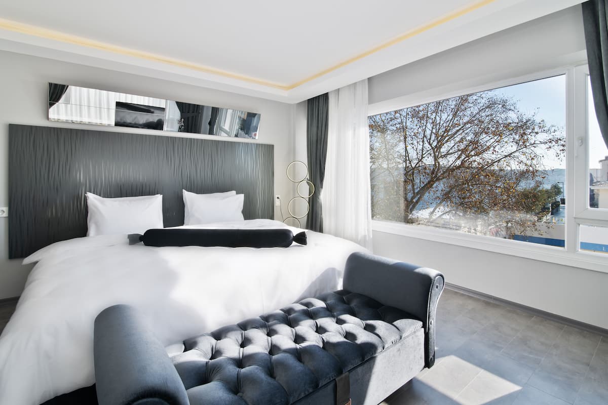 A modern hotel bedroom with a large bed, a bench, and a view of trees through the window.