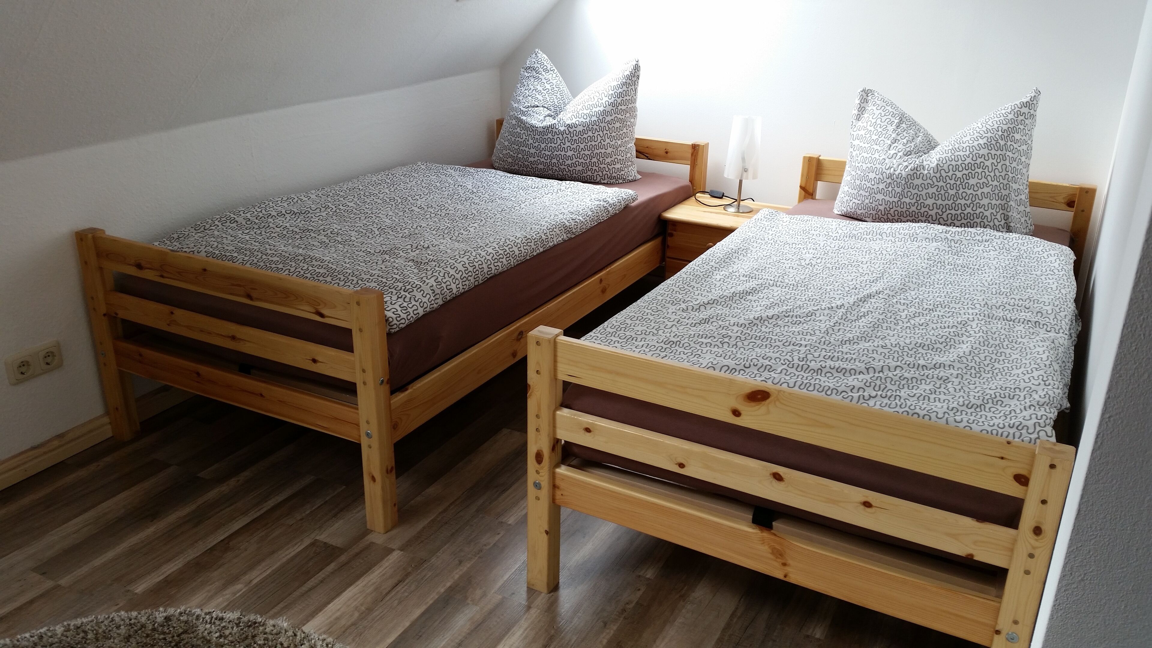 Deux lits en bois, avec des draps et des oreillers à motifs, une table de chevet avec une lampe et une prise électrique au mur.