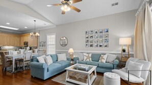 A living room with a ceiling fan, a dining area with a table and chairs, a kitchen with wooden cabinets, and a wall with framed pictures.