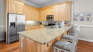 A kitchen with wooden cabinets, a stainless steel refrigerator, a microwave, granite countertops, and a bar area with bar stools.