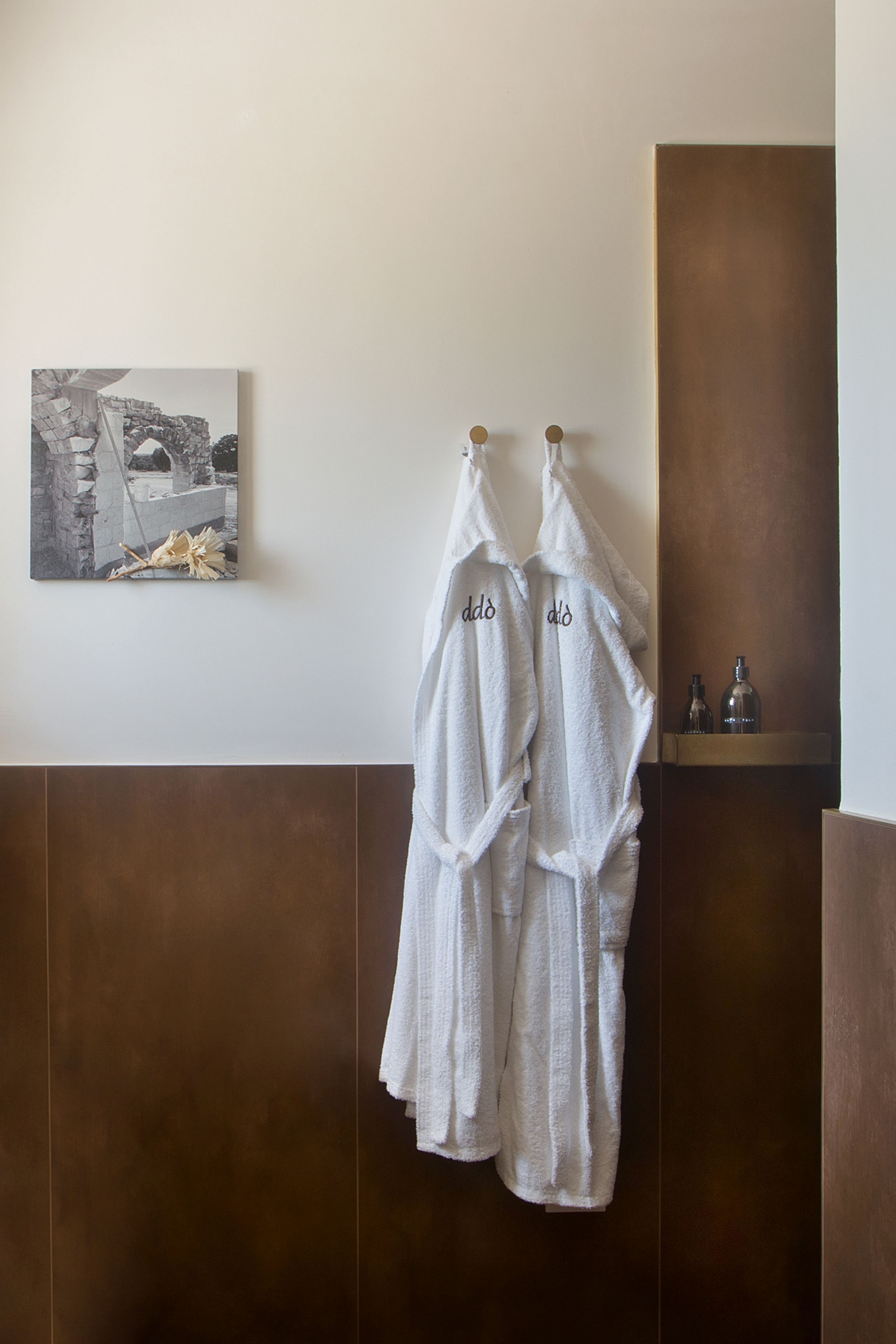 A bathroom with two white towels hanging on hooks, a small shelf with bottles, and a framed picture on the wall.