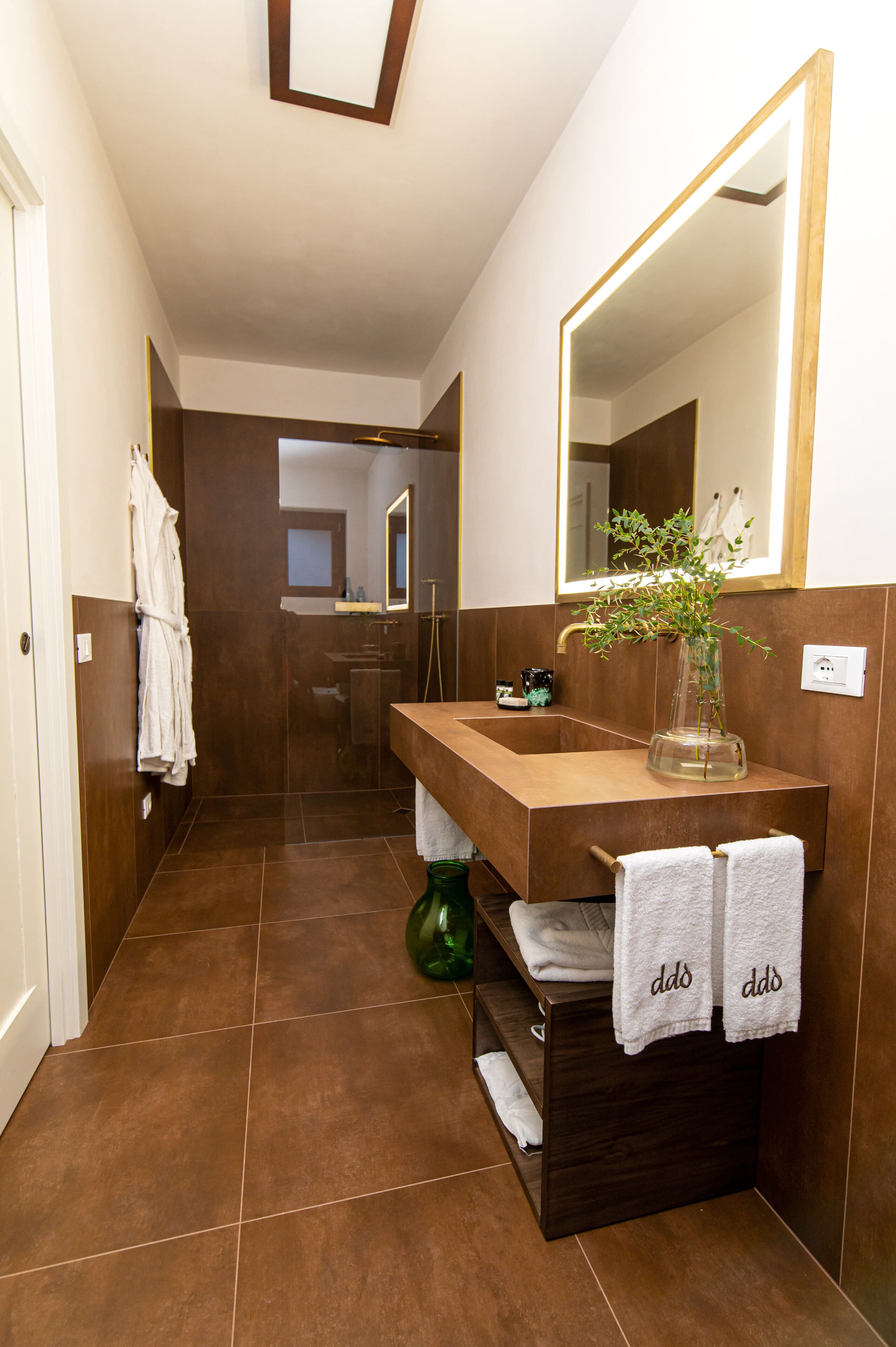 A modern bathroom with a wooden vanity, a large mirror, and a vase with green plants.