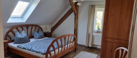 Un dormitorio con techo inclinado, una cama de madera con cabecera, un tragaluz, un armario de madera y una ventana con cortinas.