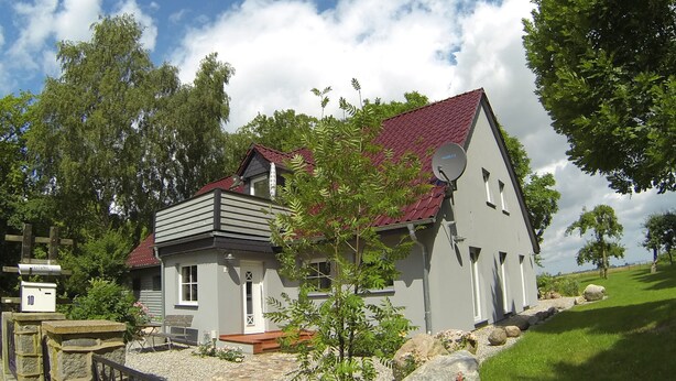 Ein zweigeschossiges Haus mit rotem Ziegeldach, einer Satellitenschüssel und einer Steinmauer mit einem Tor.