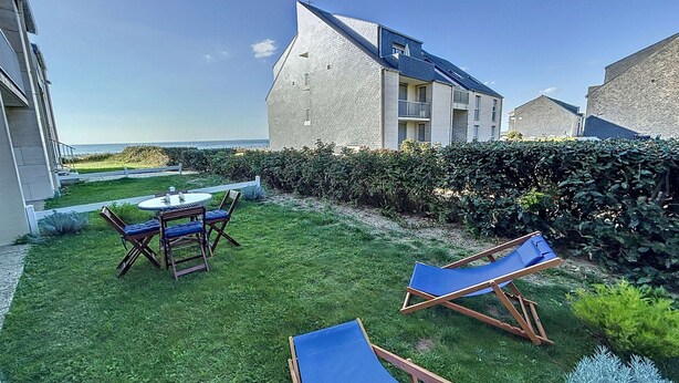 A grassy outdoor area with a table and chairs, two lounge chairs, and a building with a sloped roof.