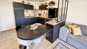 A compact kitchen with dark cabinets, a microwave, and a sink. There is a dining table with a bowl and a chair. A grey sofa with yellow pillows is in the living area.
