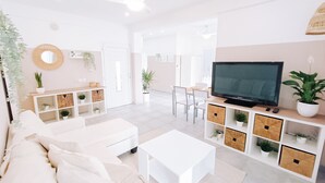 Un salon moderne avec un canapé blanc, une table basse et un téléviseur posé sur un meuble en bois. On y trouve également des plantes en pot et un meuble blanc avec des boîtes de rangement.