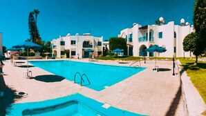 Un espace piscine avec une piscine à l’eau d’un bleu limpide, entourée de bâtiments blancs dotés de fenêtres en arc de cercle et de balcons, ainsi que de quelques arbres.