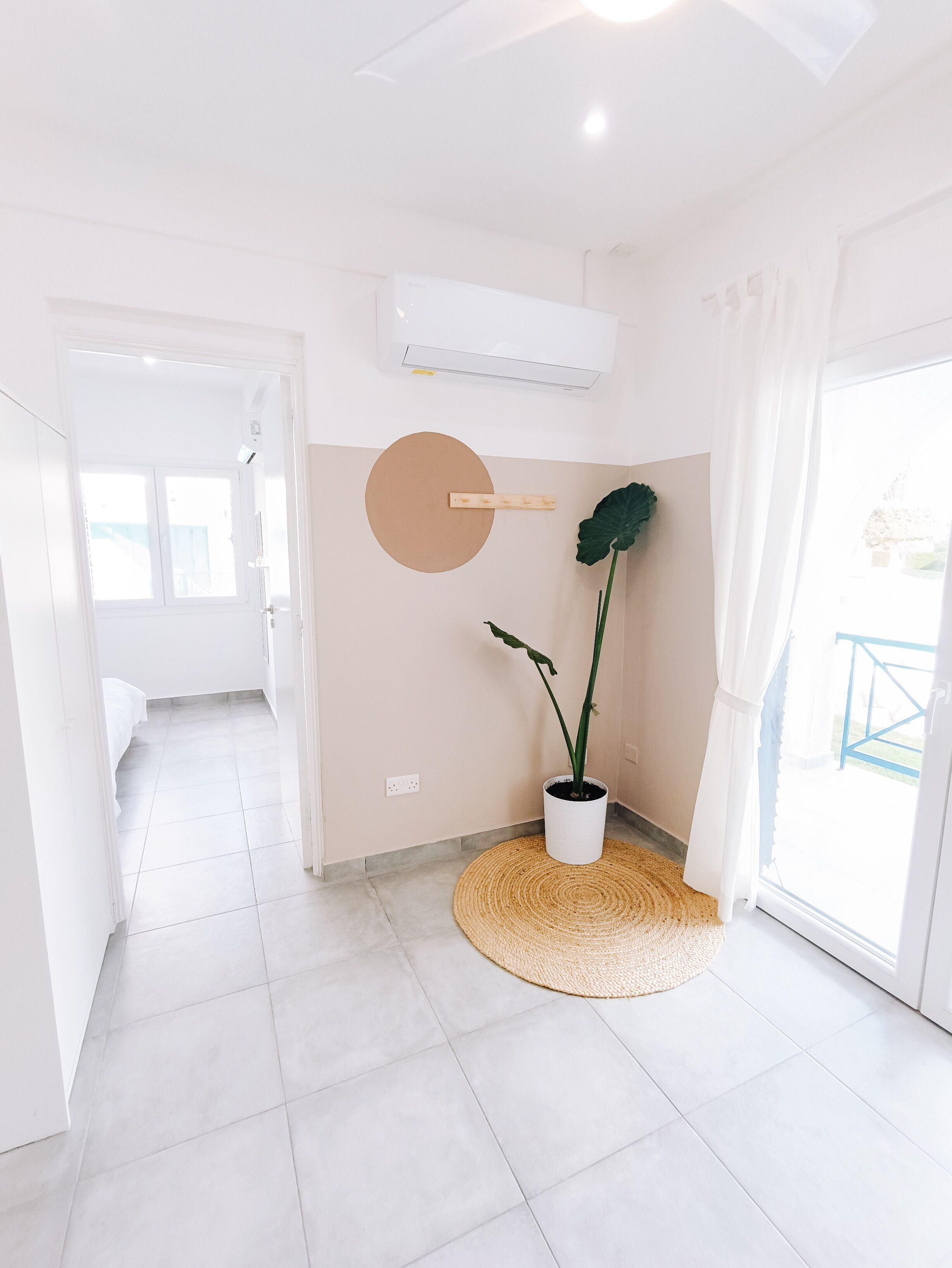 Una habitación luminosa y moderna con un ventanal grande, una planta en maceta y un tapete redondo tejido.