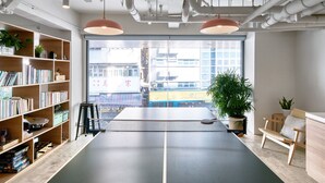 A spacious room with a ping pong table, a bookshelf, and a large window offering a view of buildings outside. - Weave Studios - Prince Edward (Kowloon)