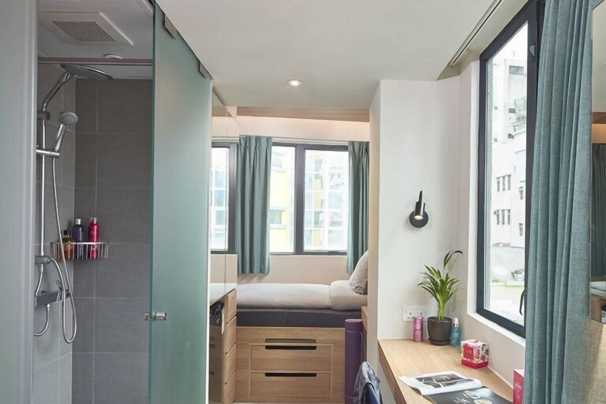 A compact hotel room with a shower, a bed, a desk, and a window with curtains.
