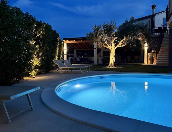 A swimming pool with a curved edge, surrounded by a paved deck. There are lounge chairs and a covered seating area with a tree in the center.