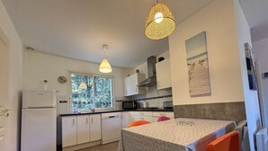 Cocina compacta con armarios blancos, un refrigerador, una ventana con vistas a la vegetación, una mesa de comedor con sillas anaranjadas y una lámpara colgante.