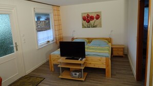 Une chambre avec un lit, un téléviseur posé sur un meuble en bois, une fenêtre avec des rideaux, un tableau représentant des tulipes rouges et un plancher en bois.