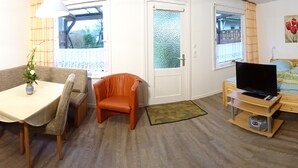 Une pièce de taille modeste comprenant une table à manger, un canapé, un fauteuil orange, un lit avec un téléviseur et une petite armoire en bois.