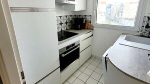 Une cuisine de taille réduite avec des meubles blancs, une cuisinière noire et une fenêtre donnant sur des immeubles.