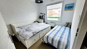 Une petite chambre bien éclairée, avec un lit simple, une fenêtre donnant sur des immeubles, un radiateur et un tableau accroché au mur.