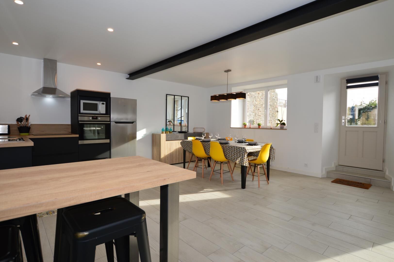 A modern kitchen with a dining area, featuring a wooden table, black bar stools, and a yellow dining chair.